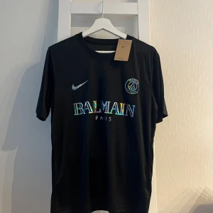 Svart PSG x Balmain fotbollströja Nike - Svart fotbollströja från Nike i samarbete med PSG och Balmain. Tröjan har korta ärmar, rund hals och är tillverkad i Dri-FIT-material som andas. Framsidan pryds av holografiskt Balmain-tryck och PSG-logga. Perfekt för dig som gillar exklusiva samarbeten.