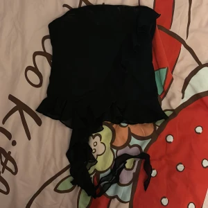 Svart asymmetrisk tubtopp med volang - Säljer en svart tubtopp med asymmetrisk nederkant och volangdetaljer. Toppen är axelbandslös och har en snygg knytning framtill som ger en cool och trendig look. Perfekt för dig som vill sticka ut lite extra.