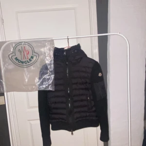 Svart Moncler pufferjacka med huva - Svart pufferjacka från Moncler med stickade ärmar och huva. Jackan har dragkedja framtill, ribbade muddar och en ficka med dragkedja på ärmen. Ikonisk Moncler-logga på vänster arm. Perfekt för dig som vill ha en snygg och varm jacka med street vibe.