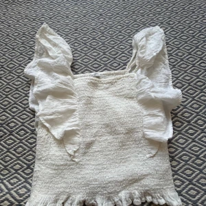 Vit volangtopp från Kappahl - Supersöt vit topp från Kappahl med smockad kropp och stora volangärmar. Toppen har en rynkad nederkant och är tillverkad i ett lätt och luftigt tyg som känns perfekt för varmare dagar. Den har en feminin och trendig look med mycket volym i ärmarna.