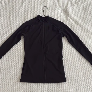 Svart ziptröja i syntetmaterial - Svart långärmad ziptröja i syntetmaterial med hel dragkedja framtill. Enkel och stilren design utan synliga loggor eller detaljer.