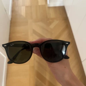 Svarta Ray-Ban Wayfarer solglasögon - Snygga svarta Wayfarer solglasögon från Ray-Ban med klassisk, lite kantig form och mörka linser. Bågarna är i blankt syntetmaterial och har Ray-Ban-loggan på sidan. Perfekt för att ge din look en cool vibe.