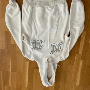 Vita mjukisbyxor med broderad logga - Snygga vita mjukisbyxor med broderade bokstäver i silver och svart på framsidan. Byxorna har resår i midjan med dragsko, sidofickor och raka ben. Perfekta för en chill och avslappnad stil.