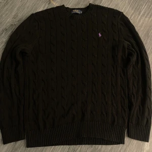 Svart kabelstickad tröja Polo Ralph Lauren - Kabelstickad svart tröja från Polo Ralph Lauren med lila broderad logga på bröstet. Klassisk rund halsringning och ribbade muddar vid ärmslut och nederkant. Perfekt för dig som gillar stilren och tidlös design.