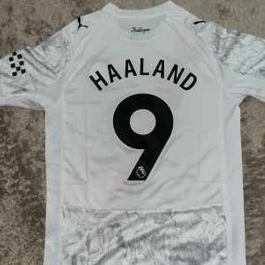 Manchester City Haaland fotbollströja Puma - Säljer en vit Manchester City fotbollströja från Puma med Haaland 9 på ryggen. Tröjan har korta ärmar, svart och röd diagonal rand på framsidan och subtilt mönster i grått. Tillverkad i återvunnet material med DryCell-teknologi. Perfekt för fans!