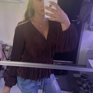 Vinröd omlottblus med snörning - Säljer en vinröd omlottblus med långa puffiga ärmar och snörning i midjan. Blusen har v-ringning och är tillverkad i ett mjukt, lätt glansigt tyg som ger en lyxig känsla. Perfekt att styla med jeans eller kjol för en trendig look.