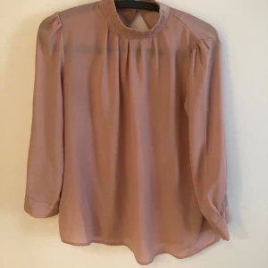 Ljusrosa blus med smockad krage - Säljer en ljusrosa blus från Wera med smockad krage och knappar i nacken. Blusen har långa ärmar med knapp vid ärmslut och är tillverkad i ett tunt, flowy material. Perfekt för dig som gillar en stilren och feminin look. Storlek 38.