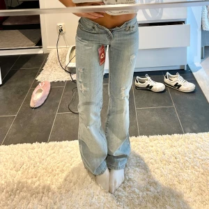 Ljusblå bootcut jeans  - Säljer ett par ljusblå jeans med bootcut passform och låg midja. Jeansen har slitna detaljer och hål framtill för en trendig look. Klassisk femficksmodell med dragkedja och knapp. Perfekta för dig som gillar en avslappnad och cool stil. Säljer då dom tyvärr är för små för mig. Midjemåttet är ca 35 cm och innerbenslängden 80 cm. Ny med prislapp