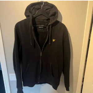   Lyle & Scott hoddie  - Svart hoodie från Lyle & Scott med gul logga på bröstet. Tröjan har dragkedja, huva med snörning och två fickor framtill. Tillverkad i mjuk bomull, perfekt för chill dagar. Långärmad och klassisk passform.