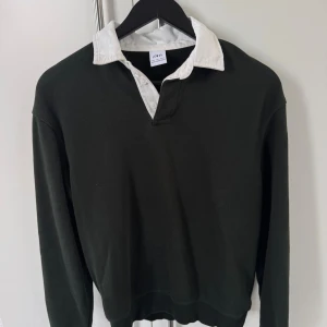 Mörkgrön pikétröja från Zara - Snygg mörkgrön pikétröja från Zara med vit krage och knappslå. Tröjan har långärmade ärmar och en klassisk, lite loose passform. Perfekt för dig som gillar preppy stil och vill ha något som sticker ut med kontrastkragen.