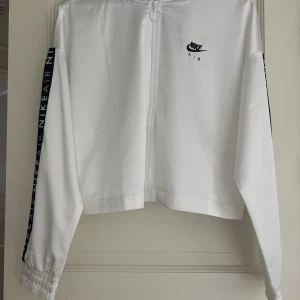 Vit Nike Air  - Snygg vit vindjacka från Nike Air med svart logga på bröstet och svarta band med texten 'NIKE AIR' längs ärmarna. Jackan har hel dragkedja framtill och elastiska muddar vid ärmslut. Perfekt för en sportig och clean look.