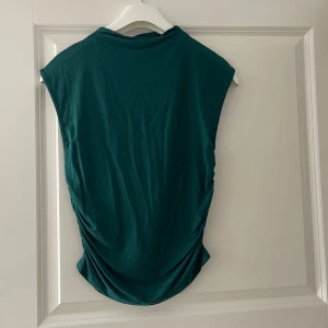 Mörkgrön ärmlös linnetopp - Snygg mörkgrön ärmlös linnetopp med lätt rynkade sidor. Toppen har en enkel och stilren design med rund halsringning och är tillverkad i mjukt material som känns skönt mot huden. Perfekt att matcha med jeans eller kjol.
