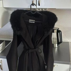 Hollies-kappa - Stilren svart kappa från hollies med lång passform, fuskpäls runt kragen och knytbälte i midjan. Jackan har långa ärmar och en klassisk siluett som ger en snygg look. Perfekt för kyliga dagar när du vill vara både varm och trendig.