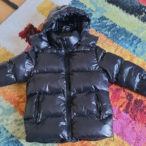 Svart glansig pufferjacka med huva XXS/XS - Säljer en svart glansig pufferjacka med huva och dragkedja framtill. Jackan har två fickor med dragkedja och en rejäl krage som skyddar mot kyla. Perfekt för kalla dagar när du vill hålla dig varm och snygg. Pris kan diskuteras