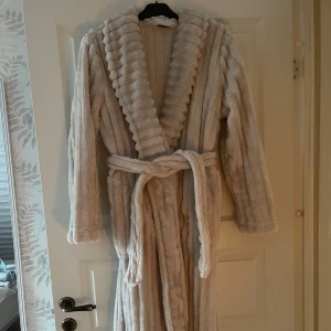 Beige fluffig morgonrock med bälte - Supermjuk och lång beige morgonrock i fluffigt material med bred krage och bälte i midjan. Har långa ärmar och ribbade detaljer längs hela plagget för extra mysfaktor. Perfekt för kalla dagar när du vill chilla hemma.
