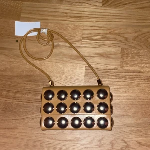 Beige axelväska med silvriga bollar - Cool beige axelväska med unika silvriga bollar på framsidan. Väskan har en slät baksida och ett smalt axelband i matchande färg. Perfekt för dig som vill sticka ut med en annorlunda accessoar. Materialet känns som syntet och designen är riktigt modern. Ny med prislapp 