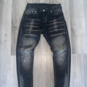 Svarta slim jeans från Dsquared2 - Svarta slim jeans från Dsquared2 med coola slitningar och färgstänk i vitt, rött och gult. Jeansen har fem fickor, smal passform och en snygg tvättad look. Röd etikett bak i midjan. Perfekta för dig som vill sticka ut med en kreativ vibe. Storlek 32