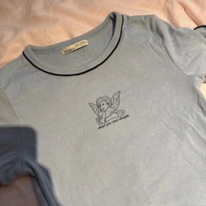 Ljusblå t-shirt med ängel från Pull&Bear - Ljusblå t-shirt från Pull&Bear med svart kant vid halsen och ärmslut. Tryck med en tecknad ängel och texten 'Angel face, devil thoughts' på bröstet. Skön passform och tillverkad i mjuk bomull. Perfekt för en chill och trendig look.