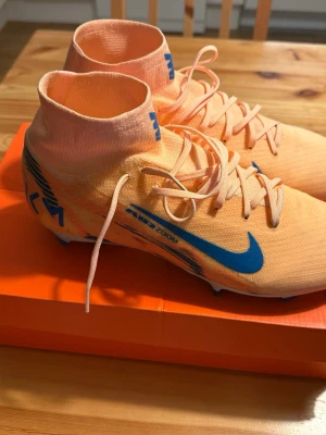 Nike Mercurial fotbollsskor orange/blå - Snygga Nike Mercurial fotbollsskor i en fräsch ljusorange färg med blå detaljer och logga. Skorna har högt skaft i strumpliknande material och snörning för bra passform. Lätta och smidiga med syntetiskt material och Air Zoom-teknologi.