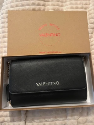 Svart handväska från Valentino - Stilren svart handväska från Mario Valentino med silverfärgad logga på framsidan. Väskan har en klassisk rektangulär form, magnetlås och kedjedetaljer på axelremmen. 