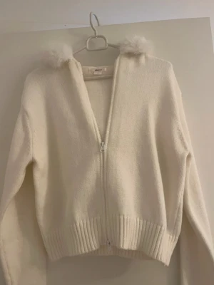 Vit stickad hoodie med fuskpäls från Gina Tricot - Vit stickad hoodie med dragkedja och huva kantad med fluffig fuskpäls. Tröjan har ribbstickade muddar och en croppad passform. 