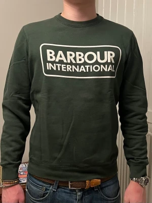 Mörkgrön sweatshirt från Barbour - Mörkgrön sweatshirt från Barbour International med stor vit logga på bröstet. Väldigt bra skick pga användning vid ett fåtal tillfällen. 