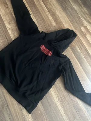 Svart Stranger Things hoodie - Säljer en svart hoodie med Stranger Things-logga i rött på bröstet. Hoodien har klassisk huva och ribbade muddar vid ärmslut och nederkant. Perfekt för dig som gillar serien och vill ha en bekväm och stilren tröja. Materialet känns mjukt och skönt mot huden.