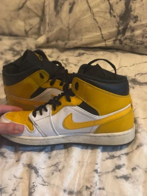 Air Jordan 1 Mid gul och vit - Snygga Air Jordan 1 Mid sneakers i gult, vitt och svart läder. Klassisk siluett med svart snörning, gul swoosh och ikonisk Jordan-logga på sidan. Perfekta för dig som vill sticka ut med färg och streetstyle.