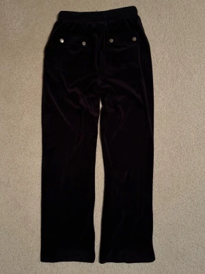 Svarta Juicy contoure velourbyxor. - Mysiga svarta mjukisbyxor i velour från Juicy Couture med raka ben, dragsko i midjan och fickor både fram och bak. Snygga silverfärgade knappar på bakfickorna och broderad logga framtill. Fint använt skick fick ett pyttelitet diskret hål på vänster framsida lår(se bild). Storlek 12-13år(ca 164cl)