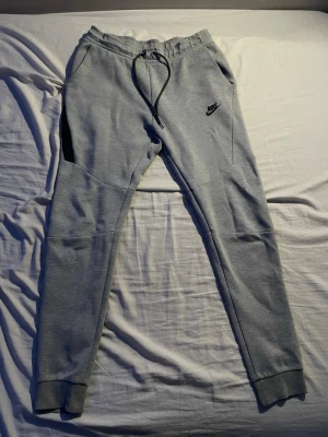 Grå mjukisbyxor från Nike med snörning - Snygga grå mjukisbyxor från Nike med svarta detaljer och snörning i midjan. Byxorna har smal passform, muddar vid bensluten och praktiska fickor. Perfekta för en avslappnad stil och riktigt sköna att ha på sig.
