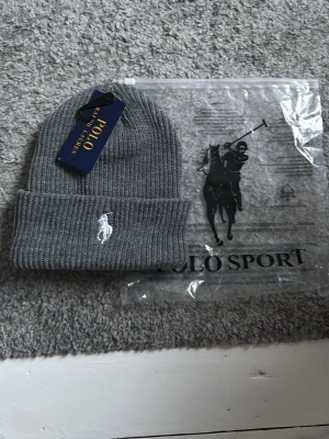 Grå ribbstickad mössa Polo Ralph Lauren - Snygg grå ribbstickad mössa från Polo Ralph Lauren med klassisk vit broderad logga framtill. Mössan har uppvikt kant och är tillverkad i mjukt material som håller dig varm och stilren under kalla dagar.
