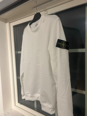Vit sweatshirt från Stone Island - osäker om den e äkta men Säljer en clean vit sweatshirt från Stone Island med klassisk rund hals och den ikoniska loggan på vänster ärm. Tröjan har en avslappnad passform och ribbade muddar vid ärmslut och nederkant. Perfekt för dig som gillar stilrena plagg med streetwear-vibe.