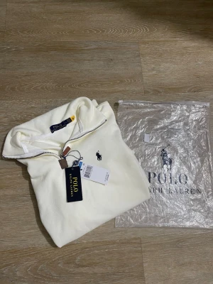Vit halvzip från Polo Ralph Lauren - Snygg vit tröja från Polo Ralph Lauren med halvzip och hög krage. Klassisk broderad logga på bröstet i mörkblått. Tillverkad i mjukt material och har långa ärmar. Perfekt för en clean och stilren look.