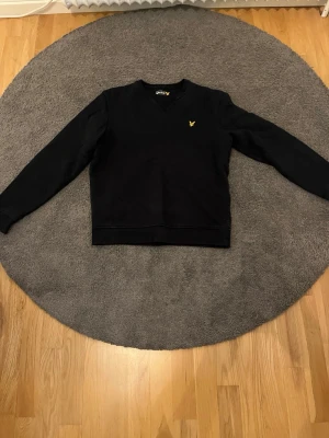 Svart sweatshirt från Lyle & Scott - Svart långärmad sweatshirt från Lyle & Scott med klassisk rund halsringning och gul logga broderad på bröstet. Enkel och stilren design som passar till det mesta. Mjuk bomullskänsla och ribbade muddar vid ärmslut och nederkant.