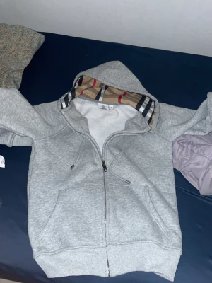 Grå hoodie med Burberry-inspirerad huva - Säljer en grå zip-hoodie från Fruit of the Loom med en huva fodrad i klassiskt Burberry-inspirerat rutmönster. Hoodien har dragkedja framtill, två fickor och justerbara snören i huvan. Tillverkad i mjukt bomullsmaterial, perfekt för chill dagar.