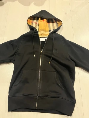 Svart Burberry hoodie - Svart hoodie med dragkedja och huva. Mjuk och skön modell med fickor framtill. Rutigt foder i huvan som ger en snygg detalj. Helt ny. Passar perfekt till vardags.