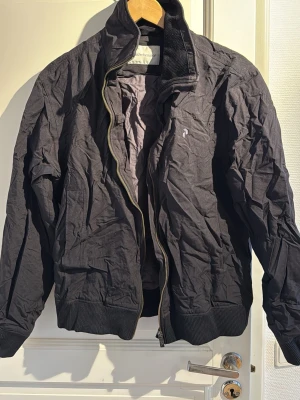 Svart bomberjacka från Peak Performance - Snygg svart bomberjacka från J.Lindeberg med klassisk ribbad krage, muddar och nederkant. Jackan har dragkedja framtill och en minimalistisk design utan synliga fickor. Materialet är tunt och lätt, perfekt för lager-på-lager och streetwear.