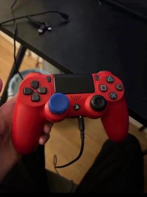 Röd PlayStation DualShock 4 handkontroll - Röd PlayStation DualShock 4 handkontroll med svart touchpad och knappar. En av spakarna har ett blått greppskydd. Trådbunden anslutning via kabel. Ergonomisk design i plast, perfekt för gaming på PS4.