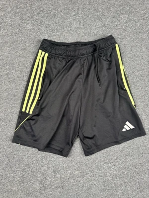 Svarta Adidas träningsshorts med snörning - Svarta träningsshorts från Adidas med gula ränder längs sidorna och snörning i midjan. Tillverkade i lätt polyester, perfekt för sport och aktivitet. Klassisk Adidas-logga framtill och AEROREADY-tryck baktill. Snygg och sportig design.