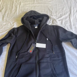 Svart hoodie från Polo Ralph Lauren - Svart hoodie från Polo Ralph Lauren med dragkedja framtill och vit broderad logga på bröstet. Tröjan har huva med snörning och två fickor framtill. Perfekt för en avslappnad stil. Tunnare material och riktigt bra skick förutom lite skadad på snörena