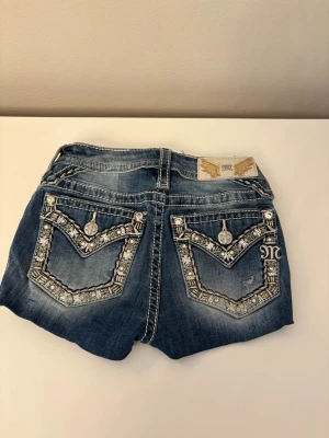 Miss me shorts  - Snygga blå jeansshorts från Miss Me med blingade detaljer och broderier på bakfickorna. Shorts med låg midja, slitningar och dekorativa knappar. Perfekta för dig som vill sticka ut med coola detaljer och unik stil. De har varit jeans som är omsydda till shorts 