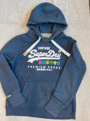 Blå Superdry hoodie med tryck - Snygg blå hoodie från Superdry med stor vit logga och färgglatt tryck på bröstet. Klassisk huva med vita snören och känguruficka framtill. Mjukt material, ribbade muddar och nederkant. Perfekt för en avslappnad streetwear-look.