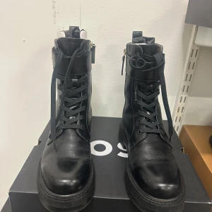 Svarta chunky kängor från Hugo boss 39  - Snygga svarta kängor i skinn från hugo boss med grov sula, snörning framtill och dragkedja på sidan. Kängorna har en cool logodetalj vid skaftet och en modern, chunky design som passar perfekt till streetwear och trendiga outfits.