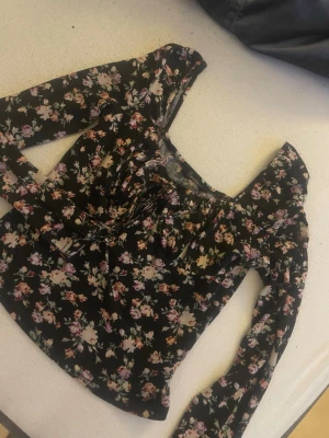 Svart blommig croppad blus - Supersöt svart blus med blommigt mönster i rosa och lila toner. Toppen har långa ärmar, croppad passform och snörning framtill vid bysten. Perfekt för dig som gillar romantisk och trendig stil.