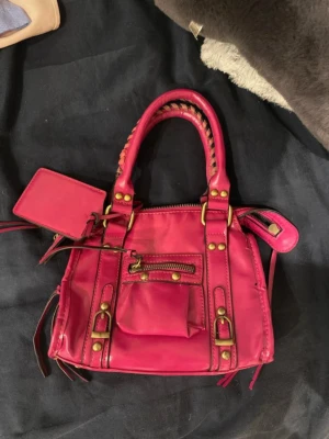 Rosa handväska med detaljer - Cool rosa handväska i skinn med guldiga nitar och dragkedjor. Väskan har två handtag med flätad detalj, en liten ficka framtill och dekorativa spännen. Perfekt för dig som vill sticka ut med en färgstark accessoar.