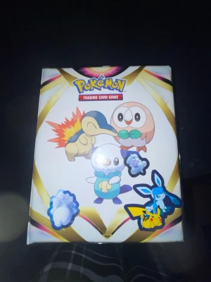 Pokémonkort med pärm (Pikatchu) - Pokémon Trading Card Game samlarkort i en pärm med Pikachu-tema. Flera kort med Pikachu och Surfing Pikachu, samt en ask med motiv av Cyndaquil, Rowlet och Oshawott. Perfekt för dig som gillar att samla Pokémonkort.