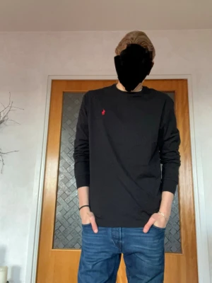 Svart långärmad topp från Ralph Lauren - Svart långärmad topp från Ralph Lauren med klassisk rund halsringning och röd broderad logga på bröstet. Tillverkad i mjuk bomull för en bekväm känsla och stilren look. Perfekt att matcha med jeans eller kjol.