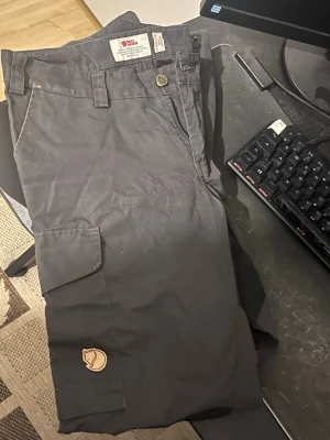 Svarta cargobyxor från Fjällräven - Snygga svarta cargobyxor från Fjällräven med klassisk logga på benet. Byxorna har flera praktiska fickor, förstärkta knän och är tillverkade i slitstarkt material. Perfekta för dig som gillar outdoor och streetwear. Storlek xs 