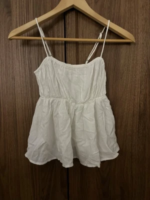 Vit somrig topp med tunna axelband - Säljer en vit, luftig topp med tunna dubbla axelband och rynkad midja. Toppen har en flowy passform och är perfekt för varma dagar. Tillverkad i ett lätt tyg som ger en avslappnad och trendig look.