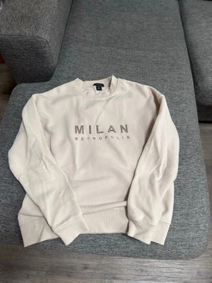 Beige sweatshirt med MILAN-tryck - Beige sweatshirt från New Yorker med texten 'MILAN METROPOLIS' broderad framtill. Tröjan har rund halsringning, långa ärmar och ribbade muddar vid ärmslut och nederkant. Storlek xs.